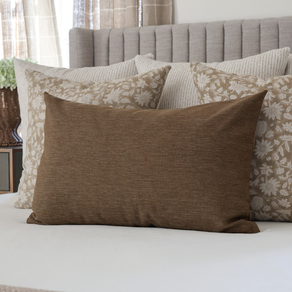 A brown textured lumbar pillow displayed on a bed with other decorative pillows and a gray tufted headboard in the background.