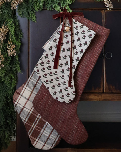 Deep berry red handmade Christmas stocking layered with plaid and floral stockings beneath evergreen garland and burgundy velvet ribbon.