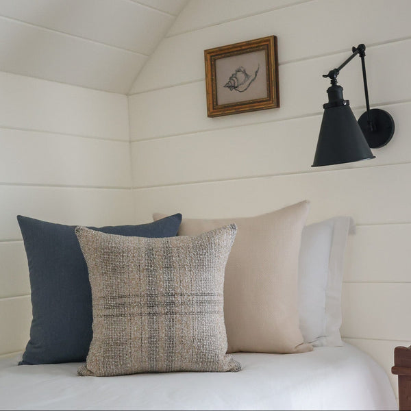 A styled bedroom corner with white shiplap walls, a black wall sconce, a small framed bird print, and a bed with white bedding and four decorative pillows, including a textured beige plaid throw pillow in the foreground.