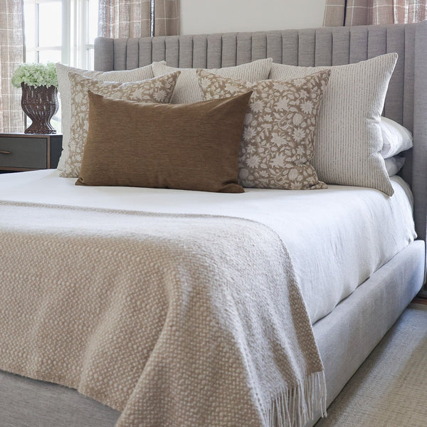 A styled bedroom scene showing a bed with a light gray upholstered headboard and footboard, dressed in white bedding with a beige textured throw blanket. Multiple pillows are arranged on the bed, including solid brown and patterned beige pillows, with plaid curtains visible in the background.