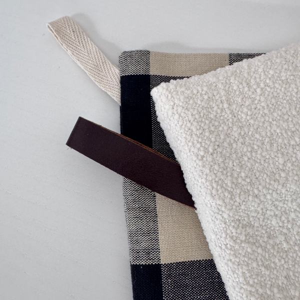A close-up of a folded textile product showing a cream bouclé side, a navy and beige buffalo plaid woven side, and a dark brown leather loop tab on a white background.