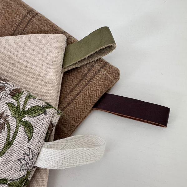 A close-up of a stack of fabric accessories, including a brown herringbone wool blanket, a beige linen cloth, a white embroidered floral textile, and a dark olive green strap, arranged on a light gray background.