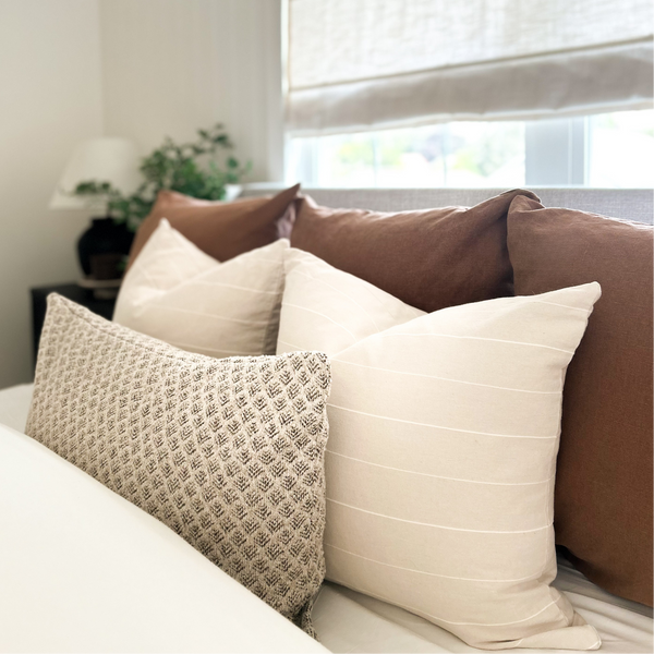 A styled bedroom scene showing a bed with layered decorative pillows in cream, beige, and brown tones, with a window and light-colored blind in the background.