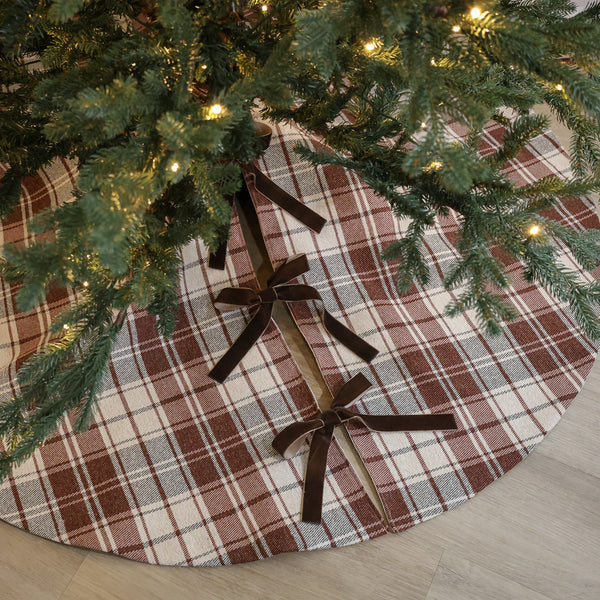 Handmade red-brown plaid Christmas tree skirt with warm cream background and brown velvet ribbon ties beneath a Christmas tree
