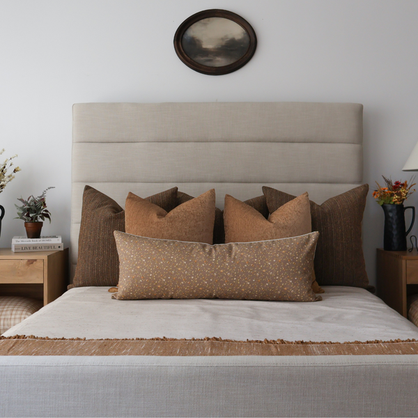 Bedroom with neutral upholstered headboard featuring layered brown and tan decorative pillows in various textures and patterns, styled on a neatly made bed with beige and rust bedding.