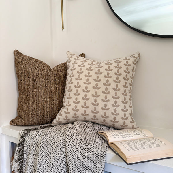 Cozy reading nook with two decorative pillows — one brown textured and one cream with tan embroidered leaves — arranged on a white bench with a patterned throw blanket and an open book beneath a round wall mirror.